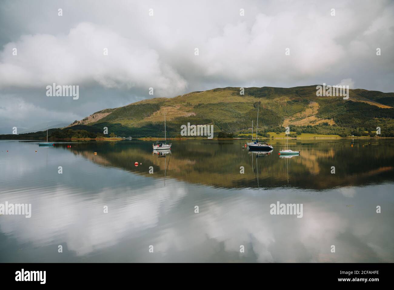 Picturesque landscape of mountain and cloudy sky reflected in tranquil water with sailboats in Glencoe on daytime Stock Photo