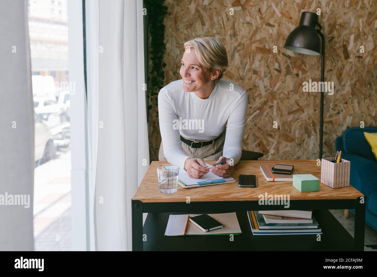 Focused adult businessWoman looking away while taking notes of plan on clipboard bending on wooden table in light office Stock Photo