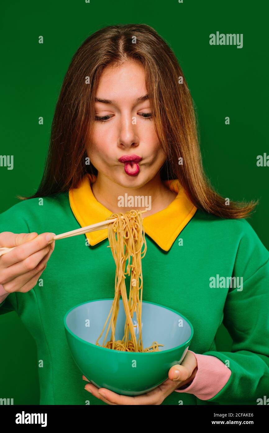 Positive young female in green shirt looking at camera grimacing making fish mouth while eating yummy instant noodles with chopsticks on green background Stock Photo