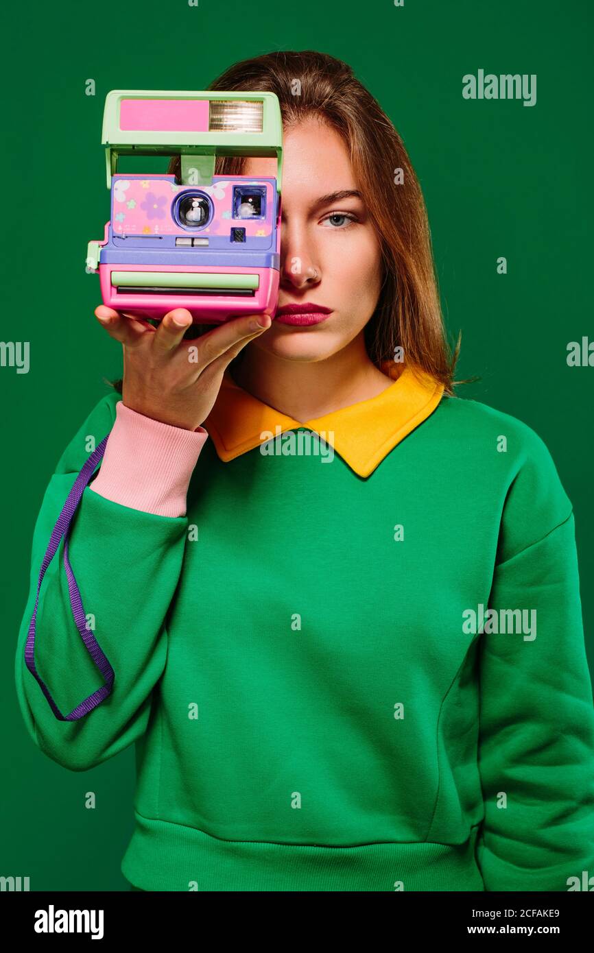 Young unemotional female in green pullover taking picture with retro instant camera while standing against green background Stock Photo