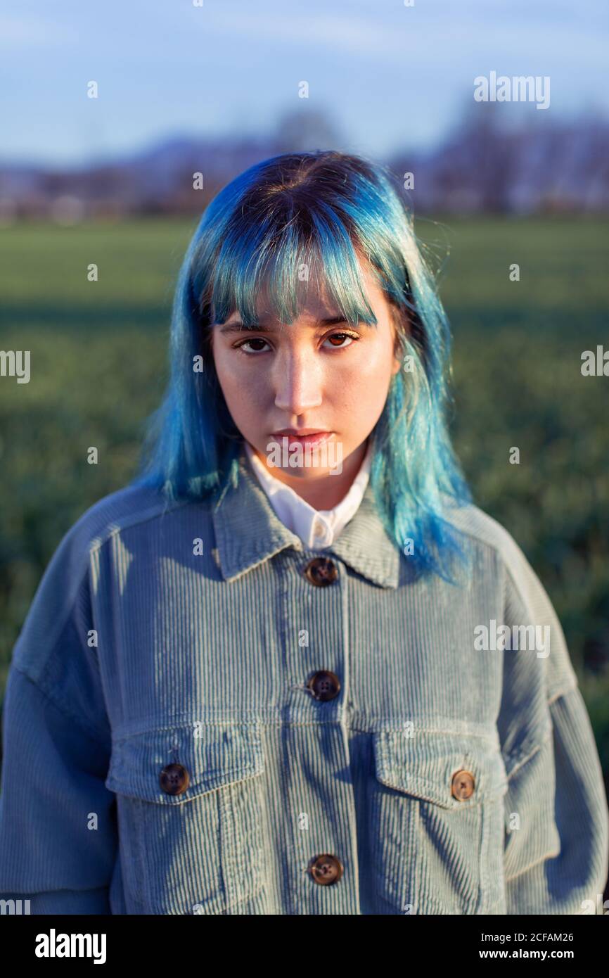 Thoughtful young female with blue hair looking at camera dressed in trendy jacket standing in green field in sunny evening Stock Photo