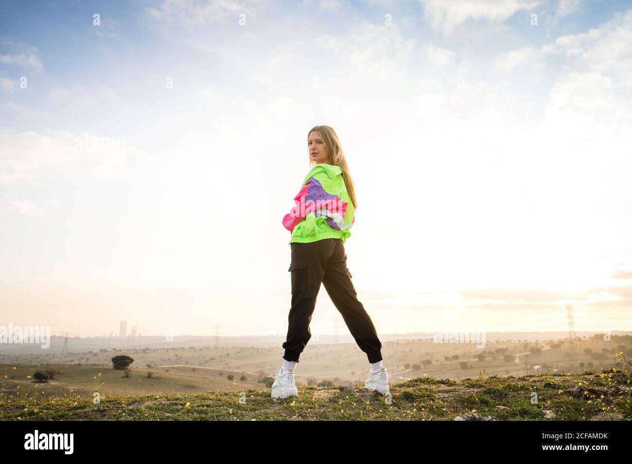 Back view of contemporary blond haired young Woman in pink bra with colorful jacket looking at camera with remote land on background Stock Photo