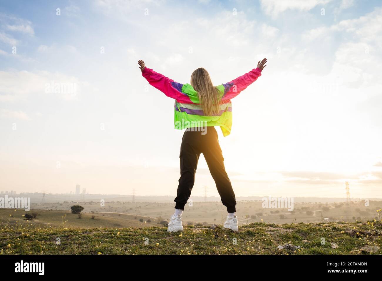 Back view of contemporary blond haired young Woman in colorful jacket raising hands with remote land on background Stock Photo