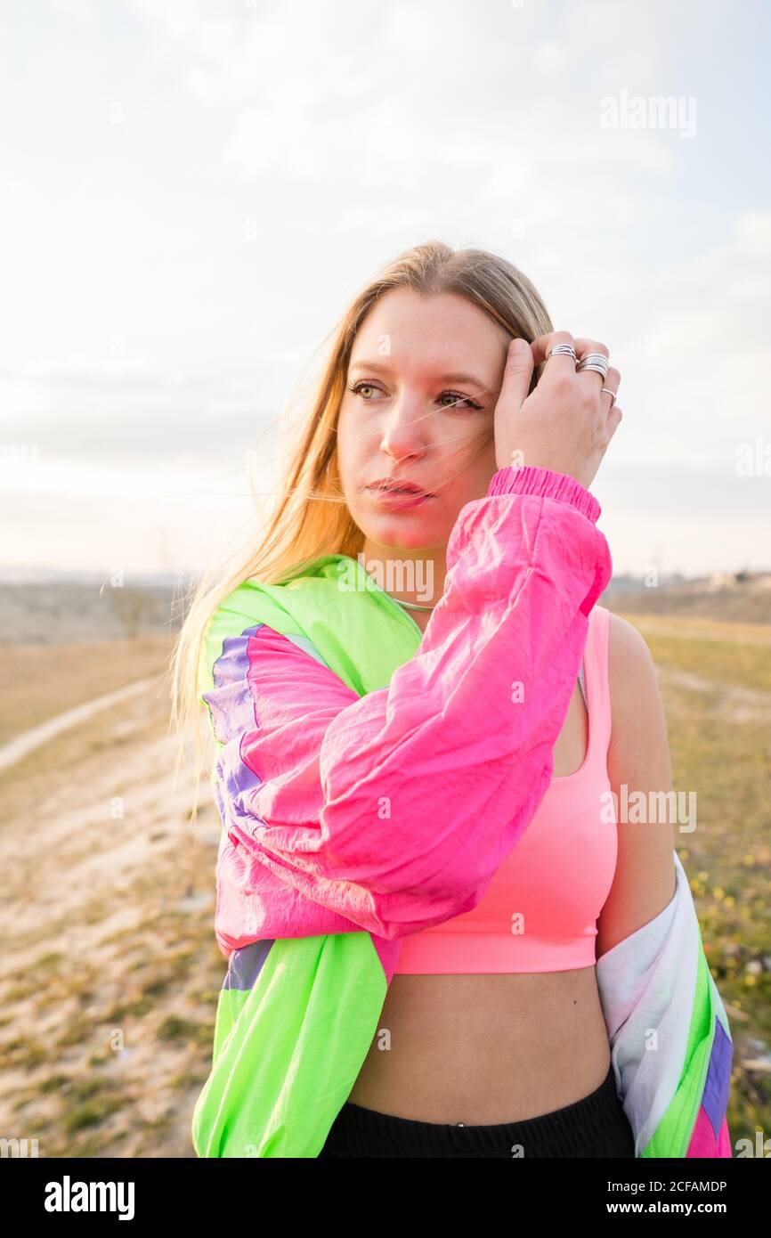 Contemporary blond haired young Woman in pink bra with colorful jacket looking away with remote land on background Stock Photo