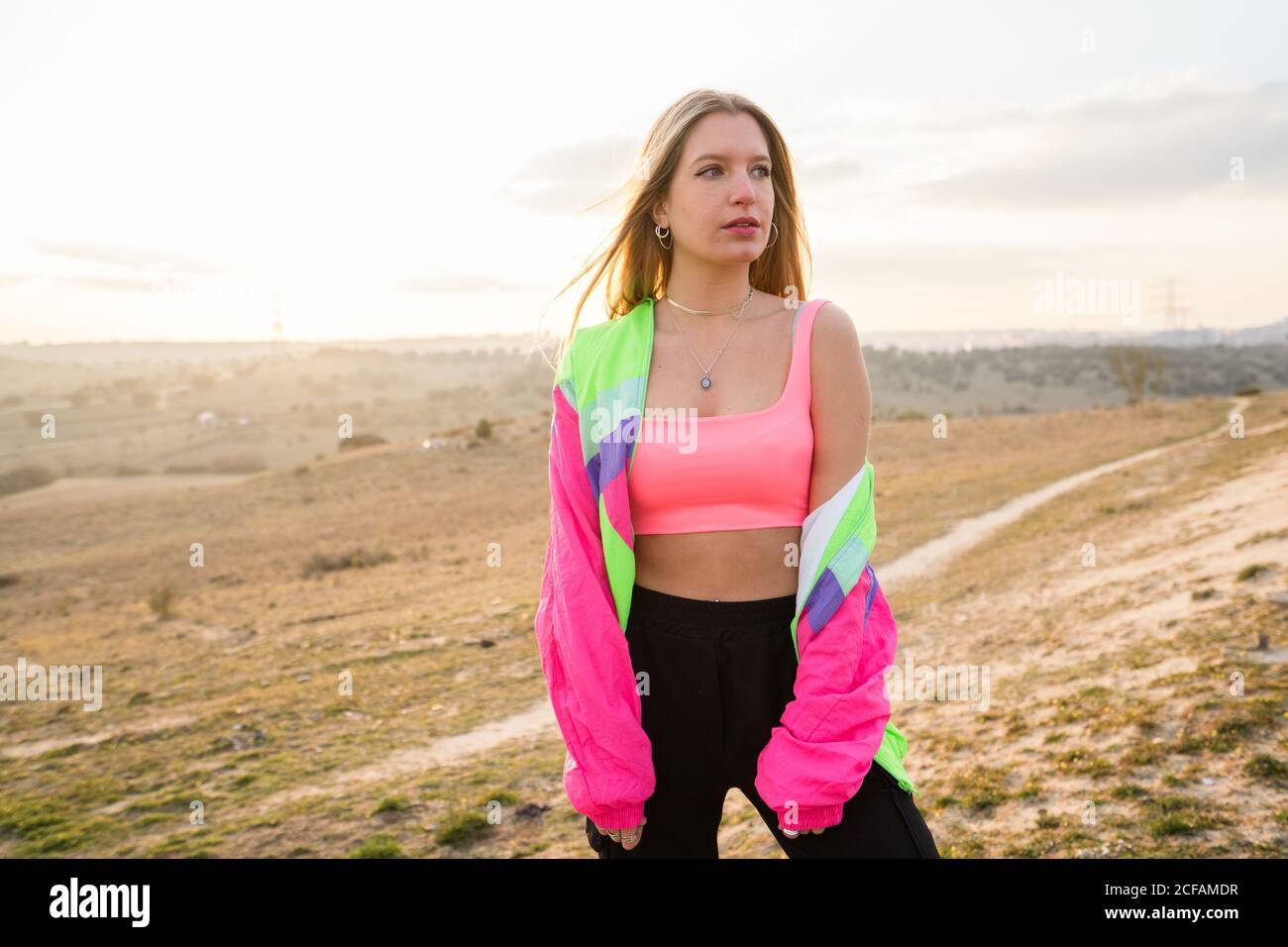 Contemporary blond haired young Woman in pink bra with colorful jacket looking away with remote land on background Stock Photo