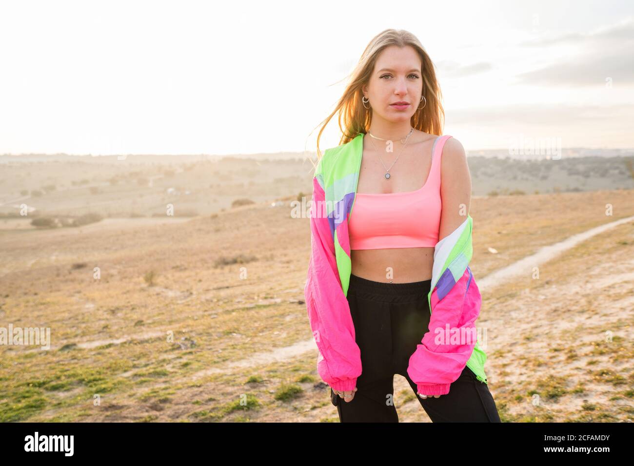 Contemporary blond haired young Woman in pink bra with colorful jacket looking at camera with remote land on background Stock Photo