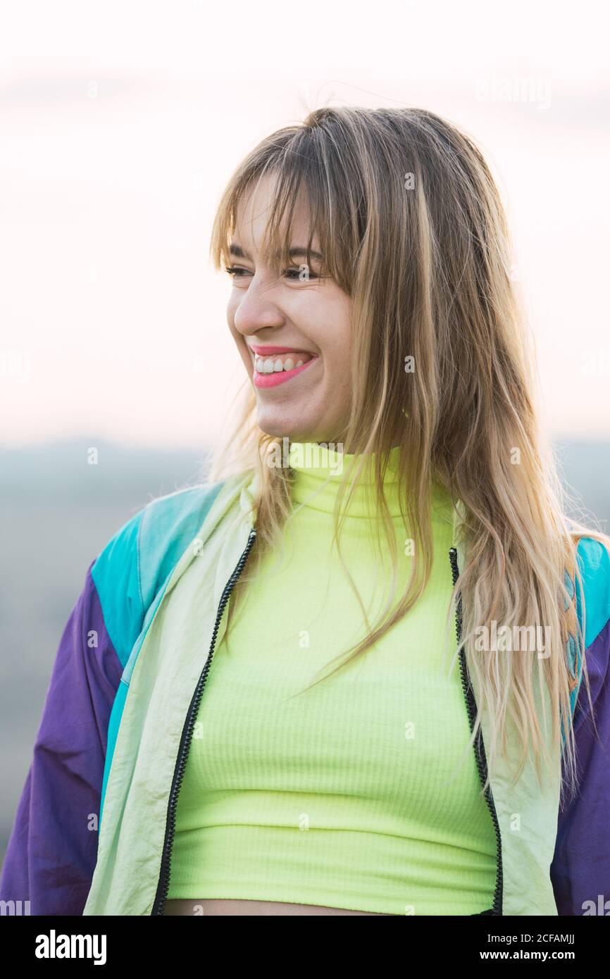 Contemporary cheerful blond haired young Woman in colorful jacket and green neon top looking away with remote land on background Stock Photo