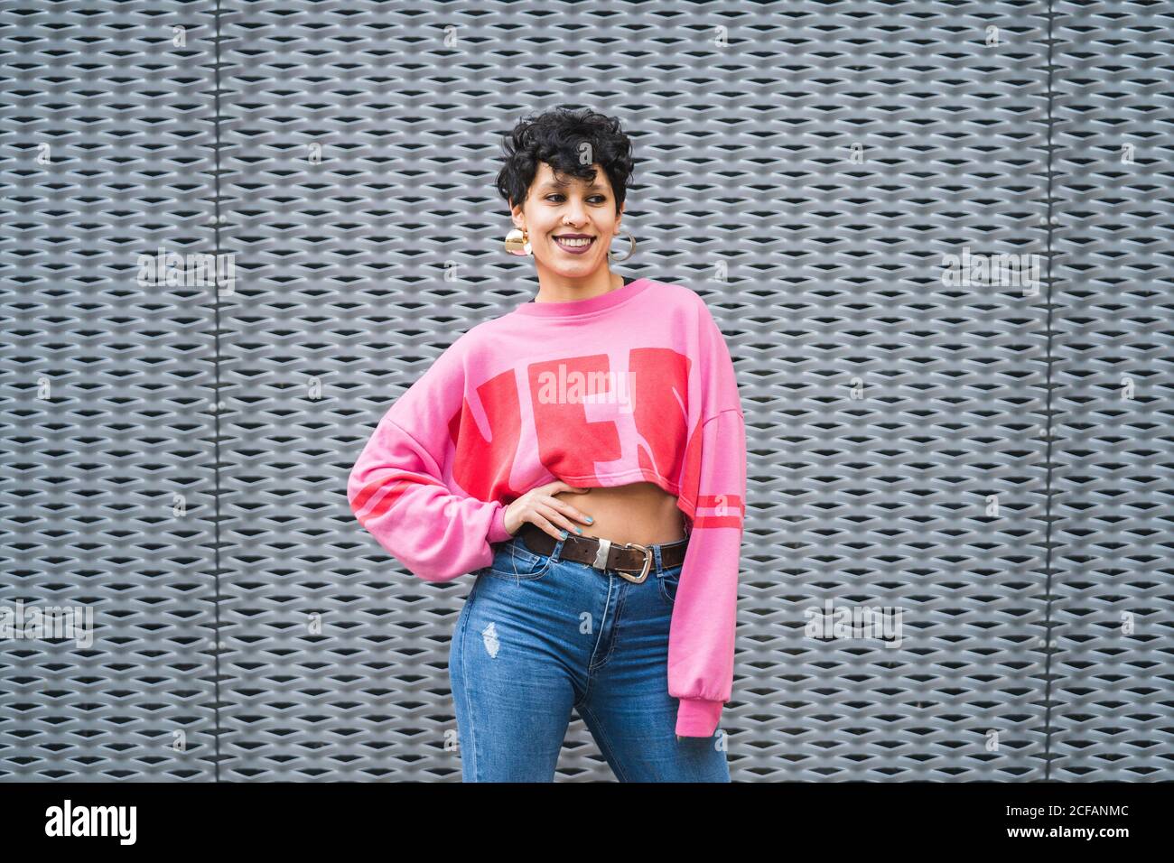 Confident cheerful ethnic black haired lady in stylish pink sweatshirt and jeans standing with hand on waist smiling away in front of grey wall Stock Photo