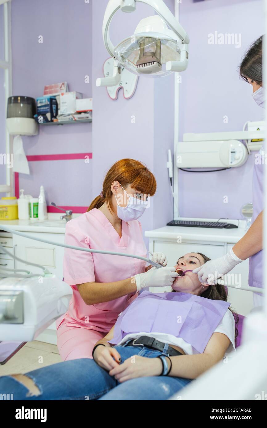 Female dentist in uniform and mask curing patient teeth with female assistant in modern dental