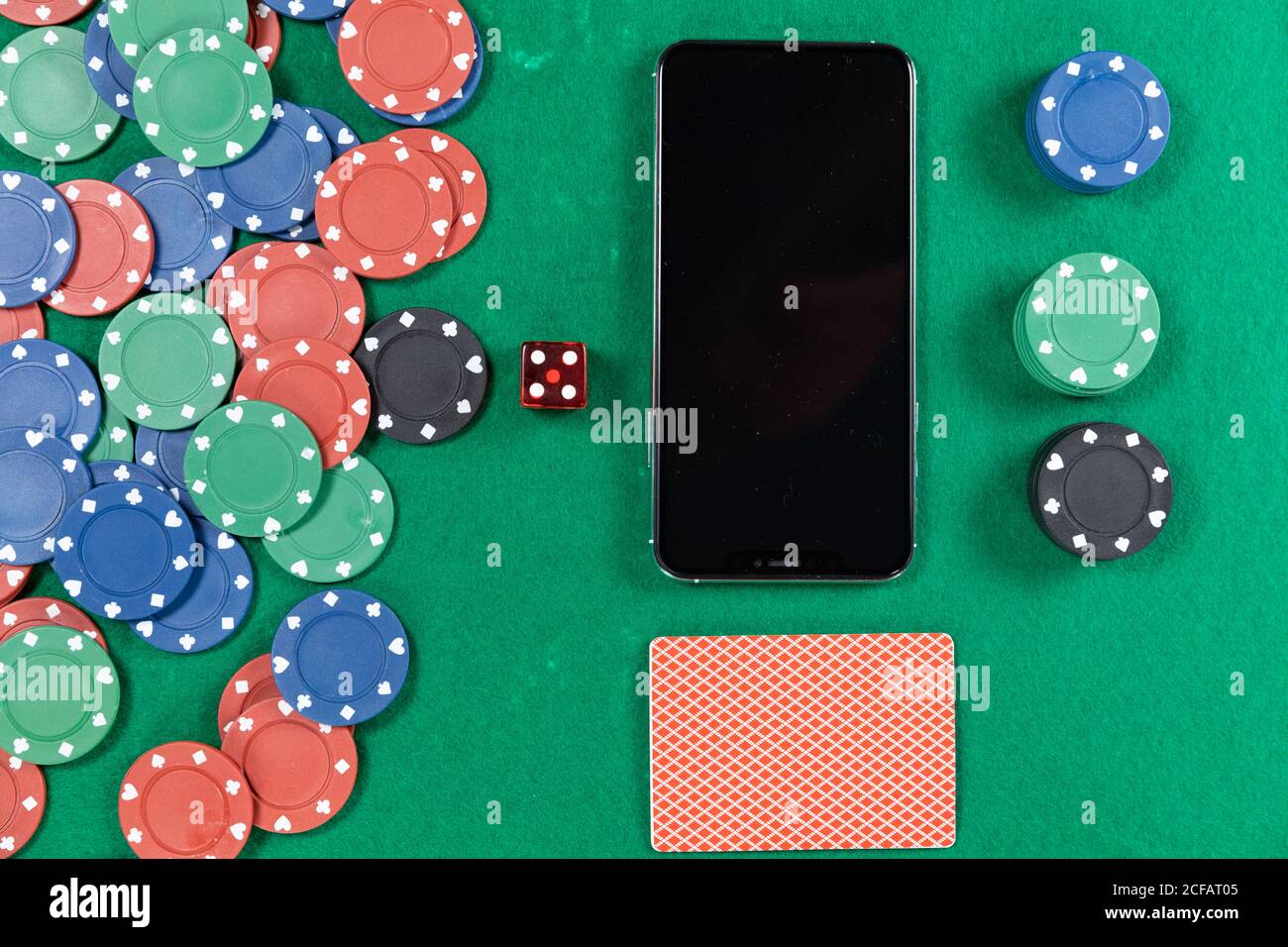 View of a black smartphone, playing cards, a red dice and colorful tokens on plain green surface Stock Photo