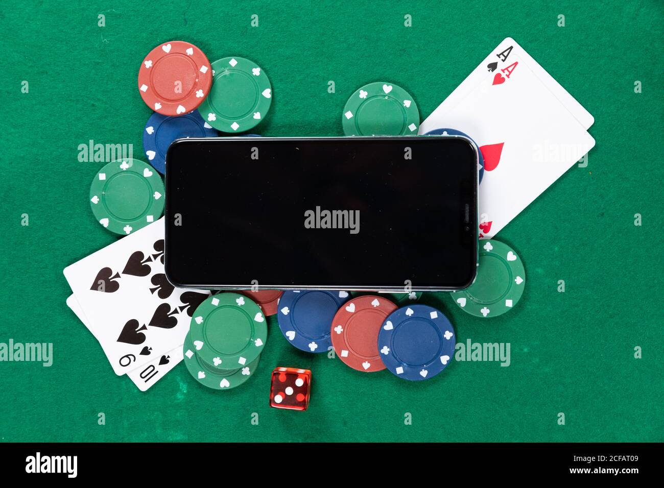 View of a black smartphone, playing cards, a red dice and colorful tokens on plain green surface Stock Photo