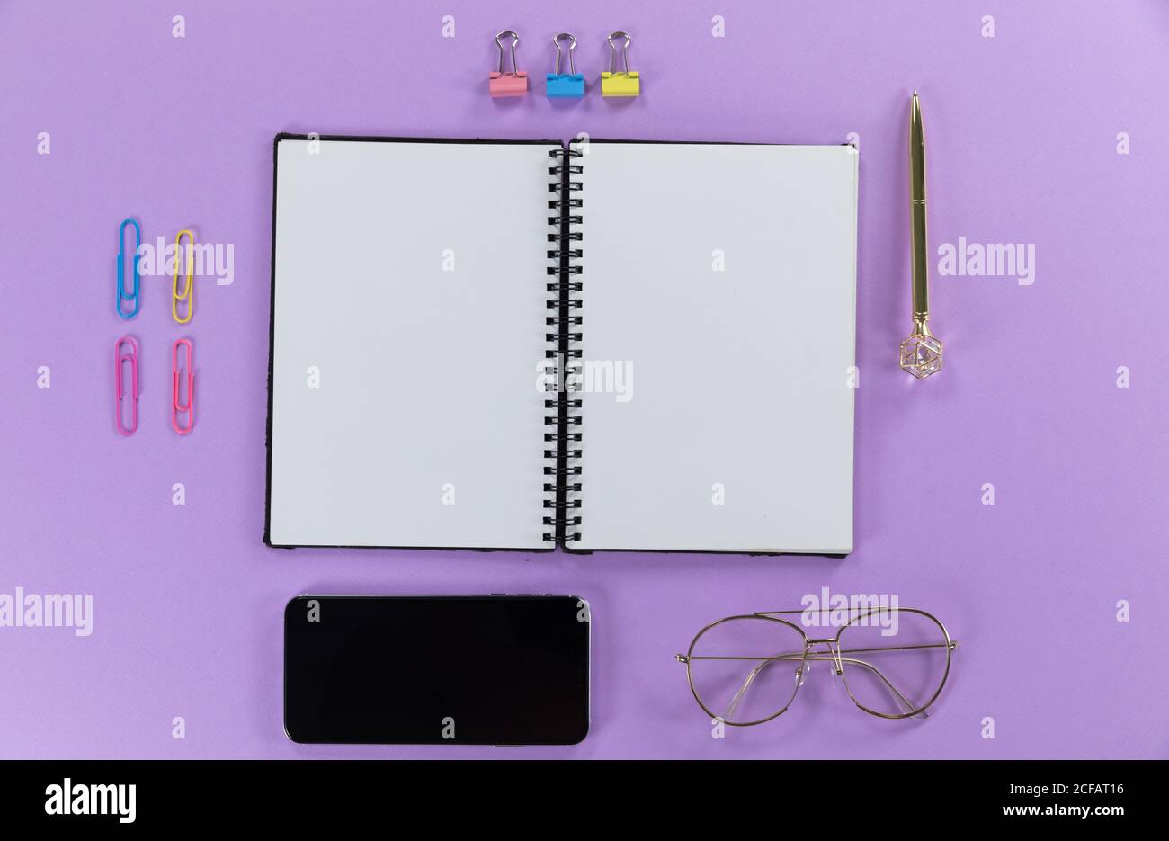 View of a black smartphone, a notebook, a pen, glasses and paperclips on plain purple background Stock Photo