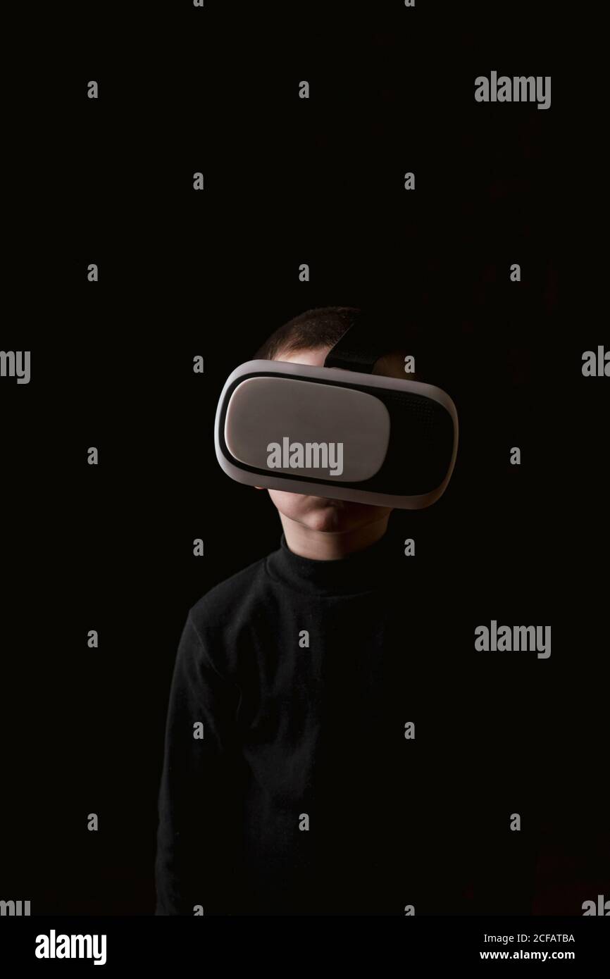 Child wearing black sweater and experiencing VR headset while standing against black wall Stock Photo