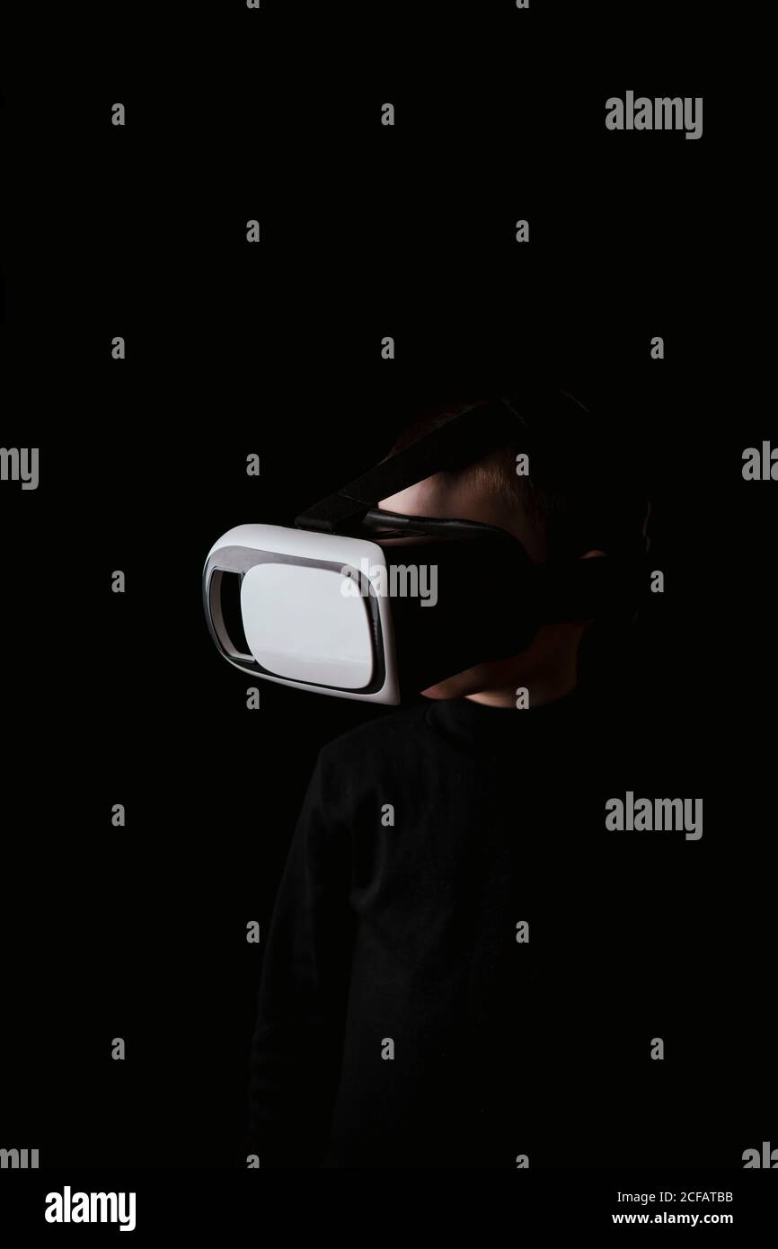 Child wearing black sweater and experiencing VR headset while standing against black wall Stock Photo