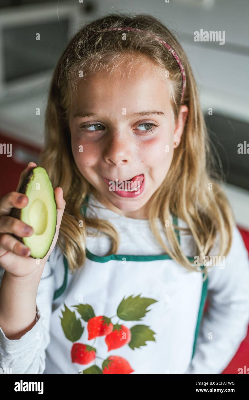 Little girl with half of tasty fresh avocado looking away and licking lips while standing in kitchen Stock Photo