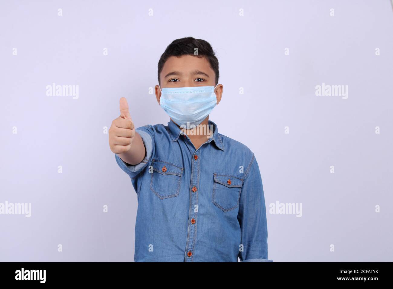 Cute Indian boy wearing surgical mask showing thumbs up or all done or like hand gesture. Isolated over white background. Stock Photo