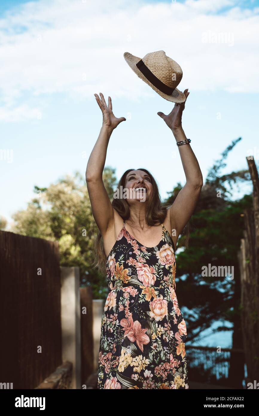 Happy adult Woman in straw hat and floral dress smiling on sunny street Stock Photo