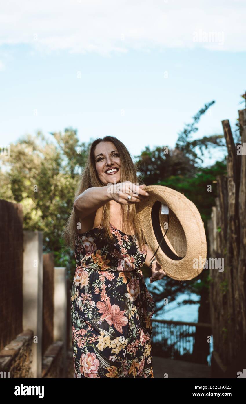 Happy adult Woman in straw hat and floral dress smiling at camera on sunny street Stock Photo