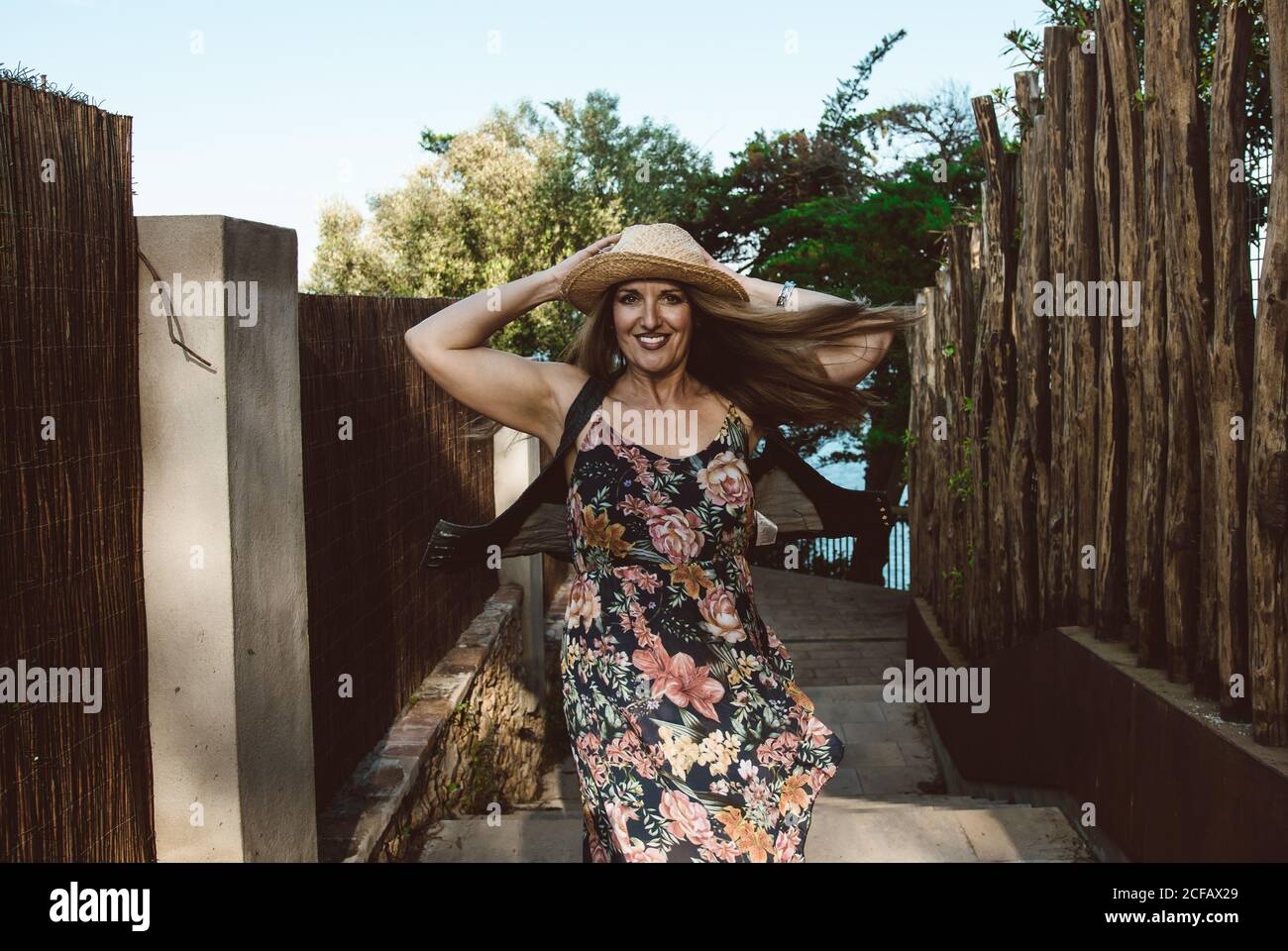 Happy adult Woman in straw hat and floral dress spinning around smiling at camera on sunny street Stock Photo