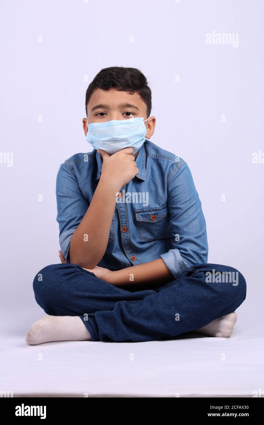 indian cute young boy wearing surgical mask pointing finger towards top while seating on floor. Stock Photo