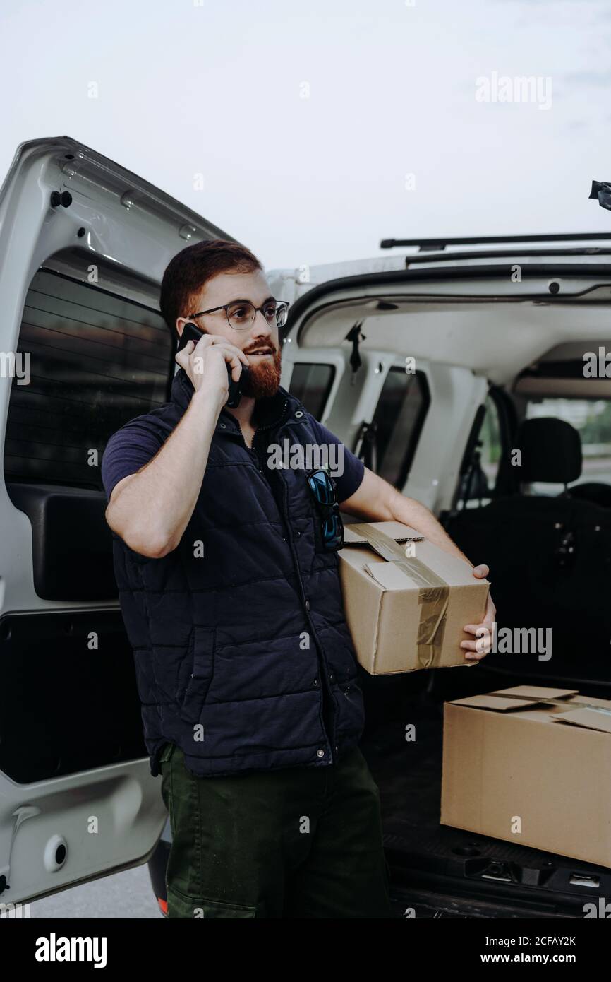 Pleased adult bearded courier in glasses holding boxes and making warning call to customer for further delivery while standing near car trunk and looking away during daytime Stock Photo