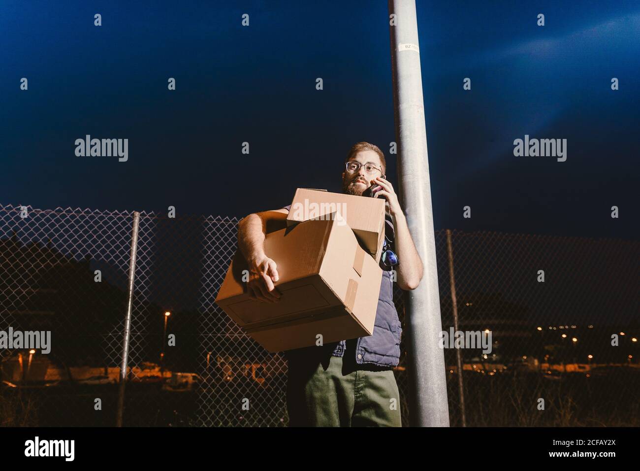 Calm pensive tired adult bearded courier in glasses with boxes making warning call to customer for further delivery and looking away while standing and leaning on streetlight post on street in evening Stock Photo