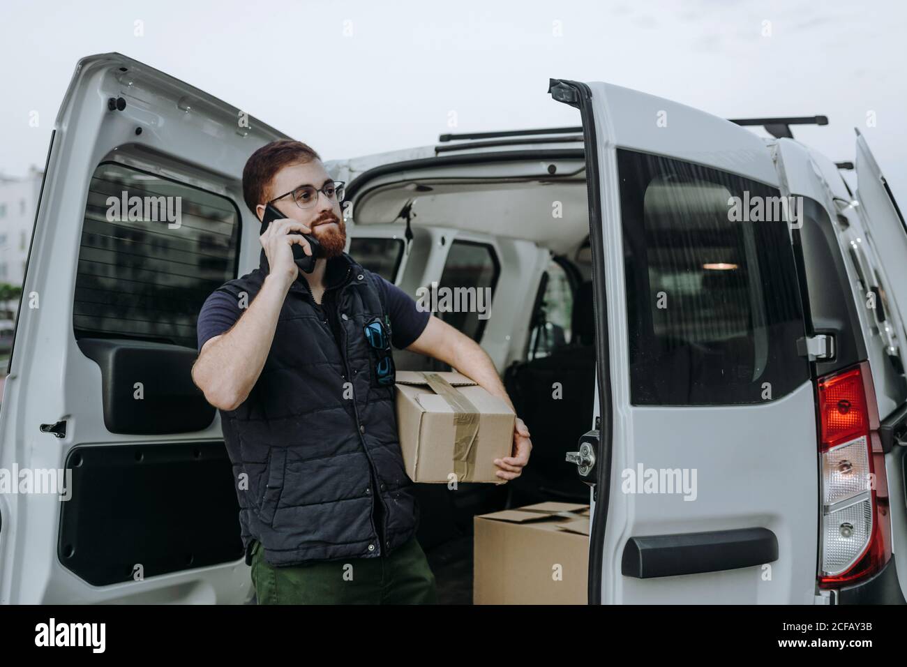 Pleased adult bearded courier in glasses holding boxes and making warning call to customer for further delivery while standing near car trunk and looking away during daytime Stock Photo