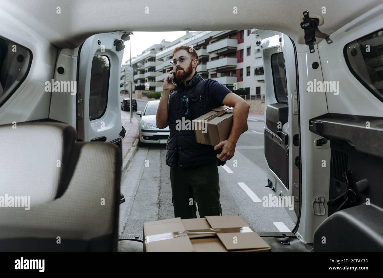 Pleased adult bearded courier in glasses holding boxes and making warning call to customer for further delivery while standing near car trunk and looking away during daytime Stock Photo