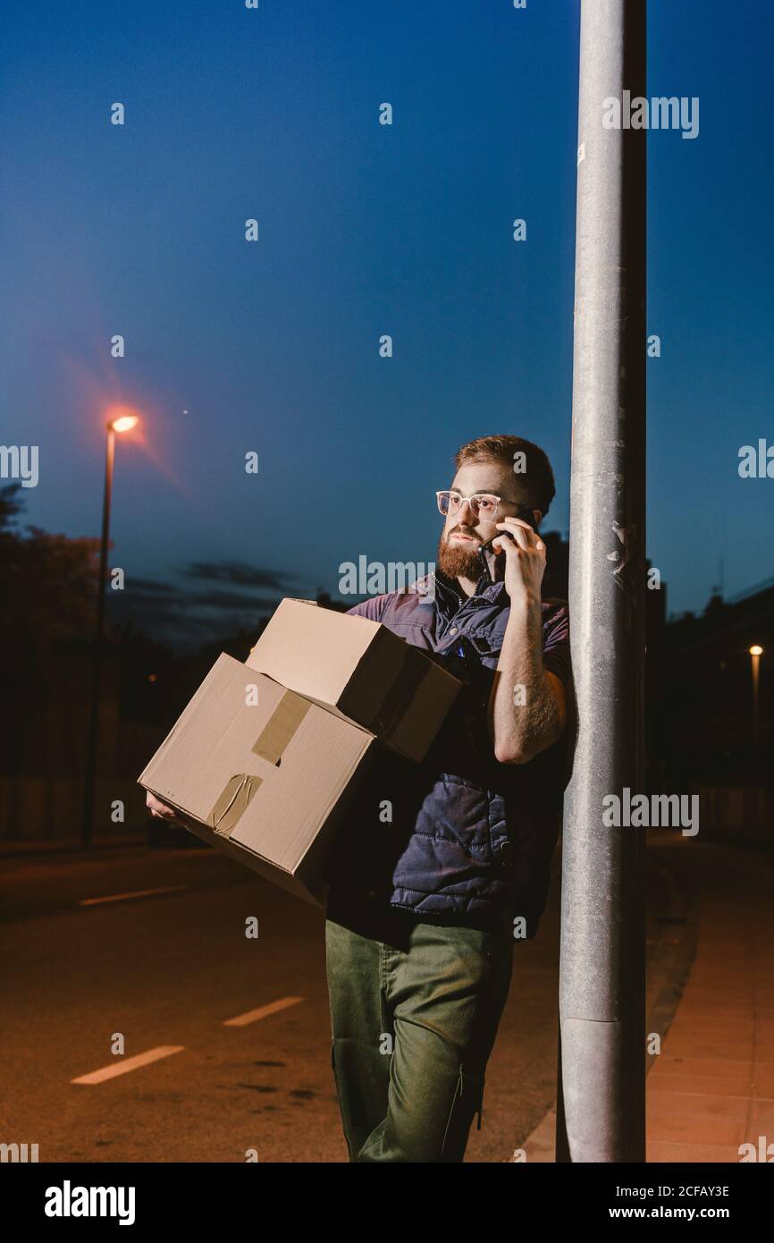 Calm pensive tired adult bearded courier in glasses with boxes making warning call to customer for further delivery and looking away while standing and leaning on streetlight post on street in evening Stock Photo