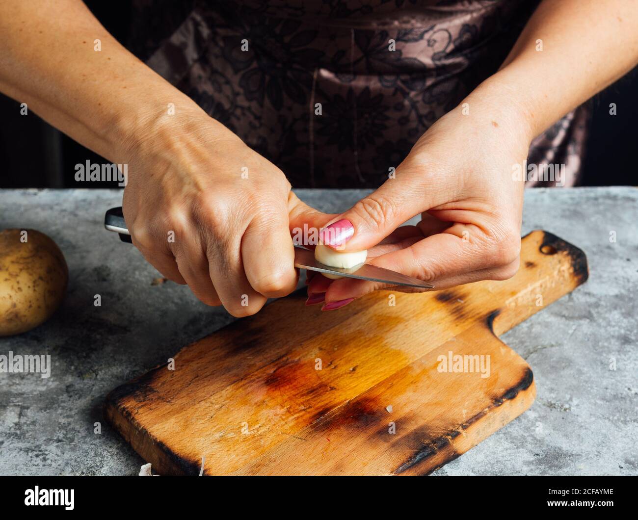 unrecognizable female cook cutting clove of garlic with knife while standing at table with wooden cutting board and preparing healthy dinner at home Stock Photo