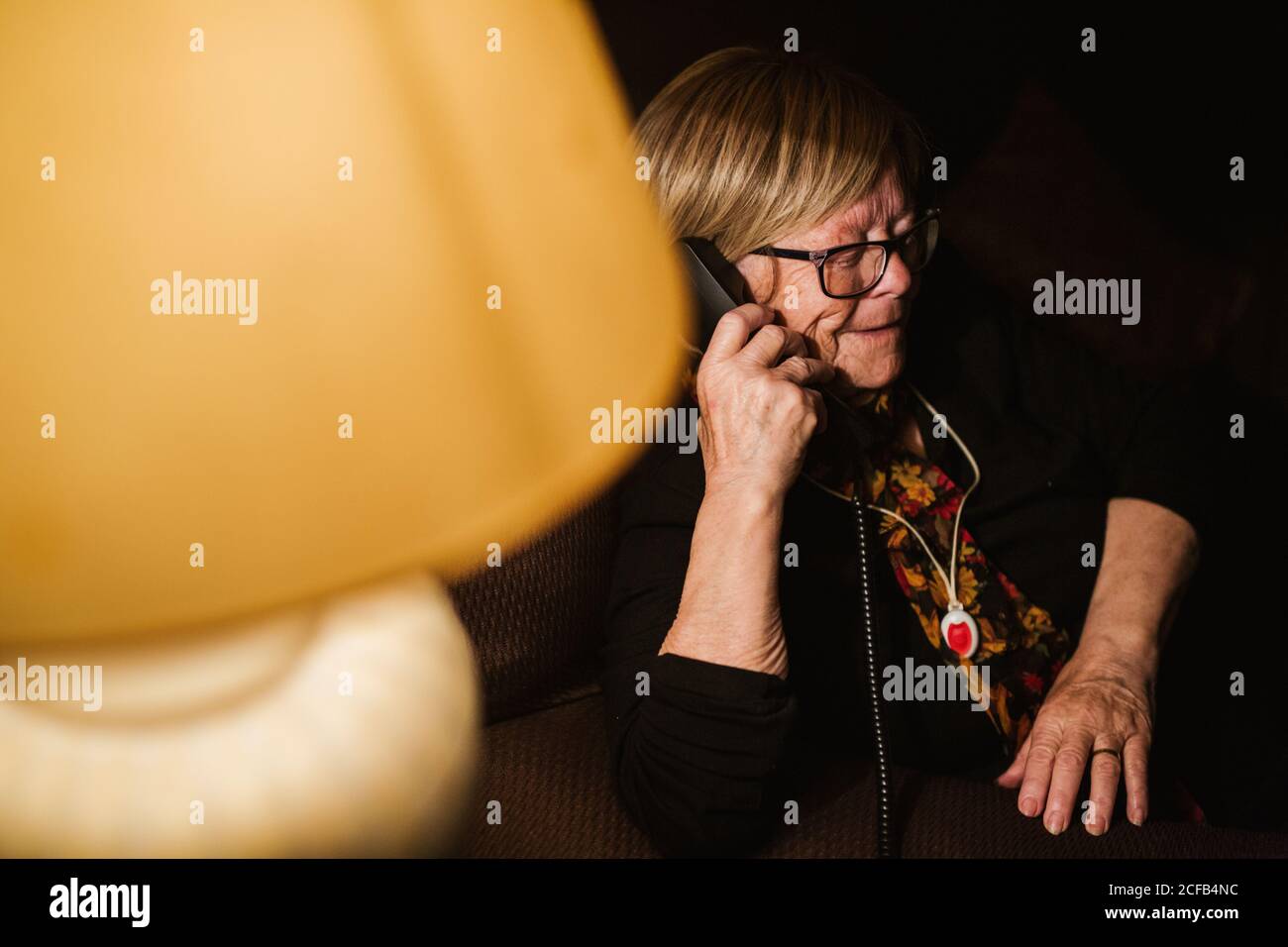 Glad elderly lady smiling and answering phone call while sitting in dark room in evening at home Stock Photo