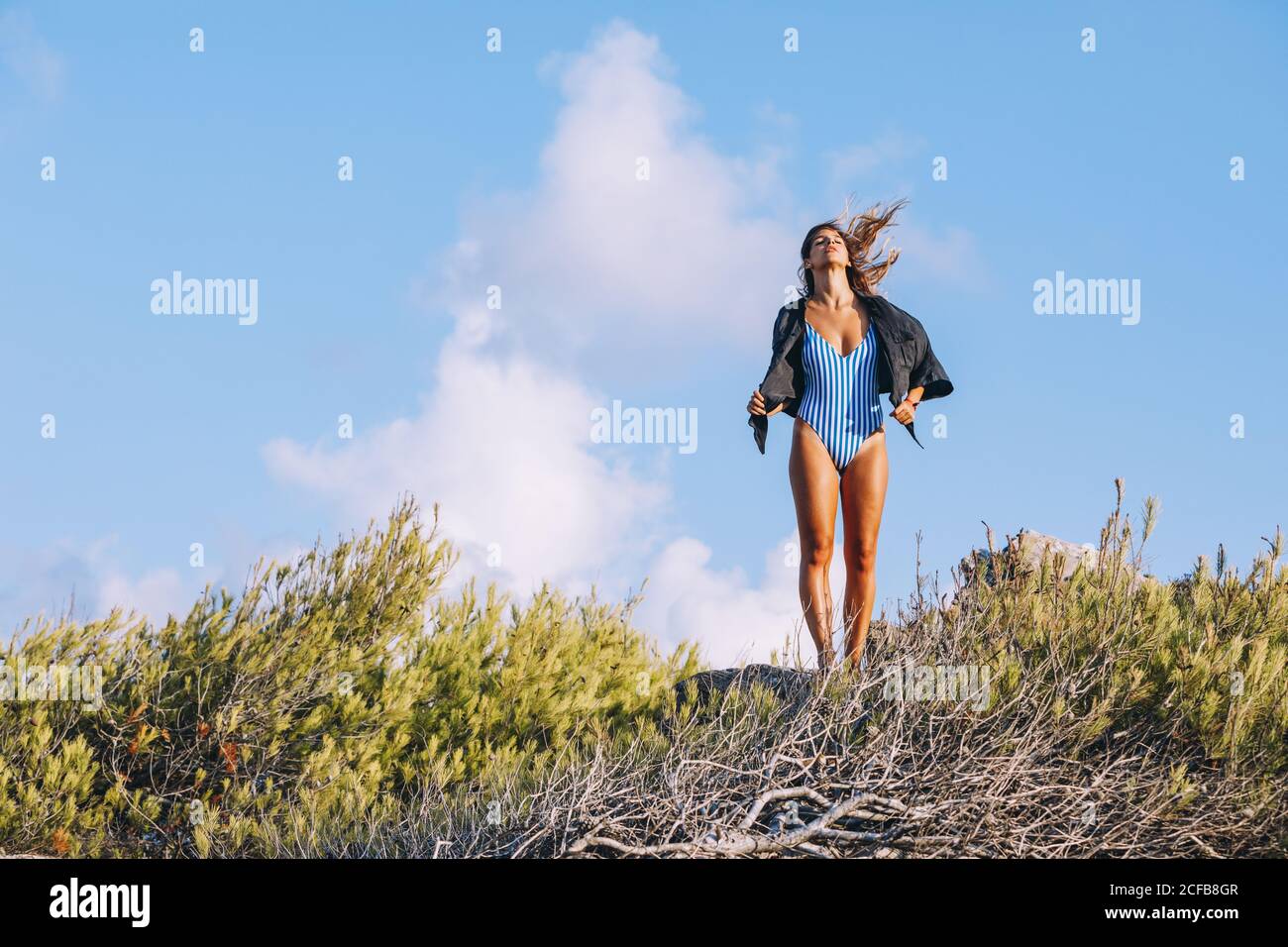 Contended tanned lady in trendy striped swimsuit and shirt on shoulders relishing in weather while walking on countryside with blue sky on background Stock Photo