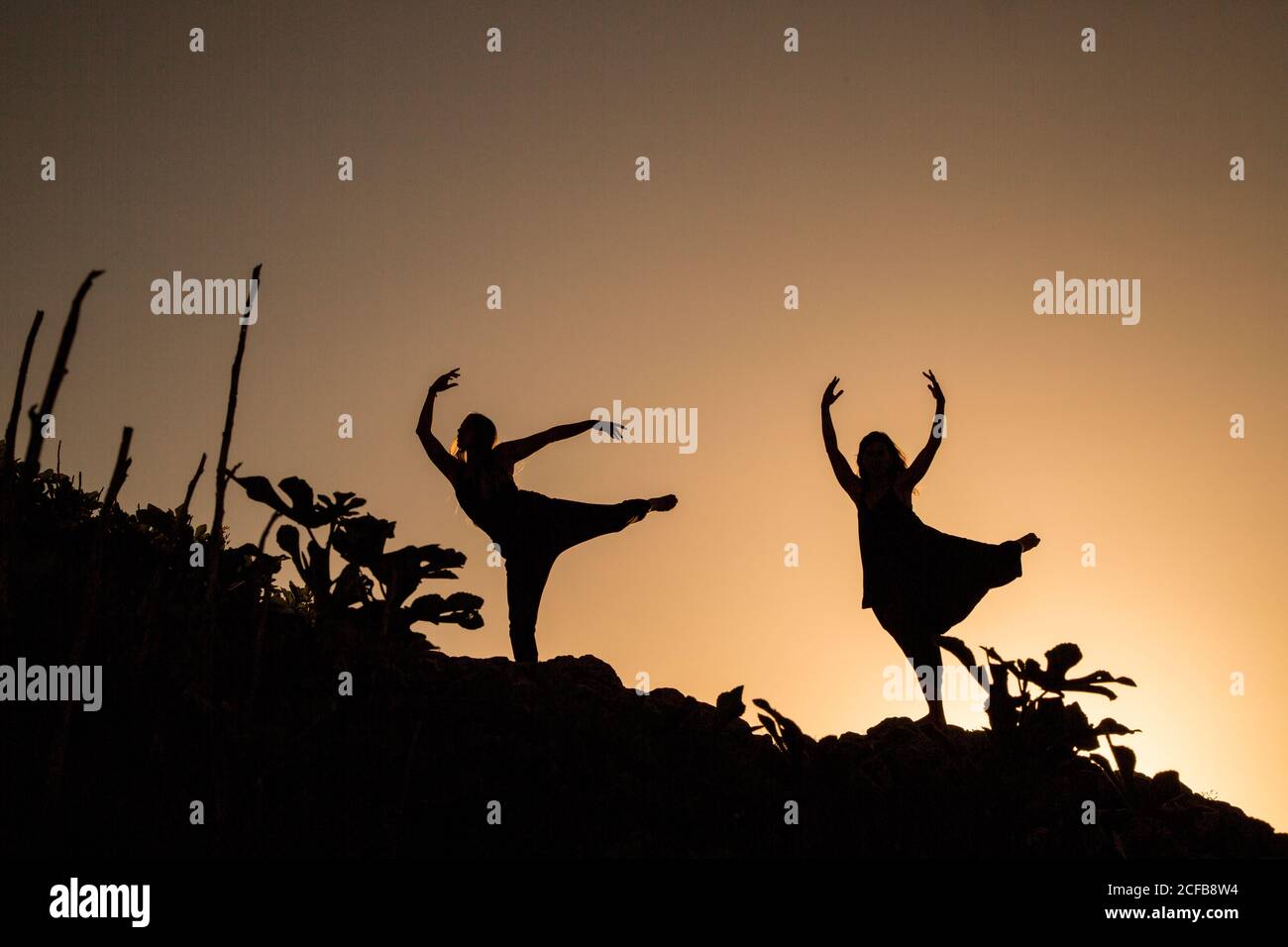 Young ballerinas with upped legs and hands dancing on hill in obscurity at sunset Stock Photo