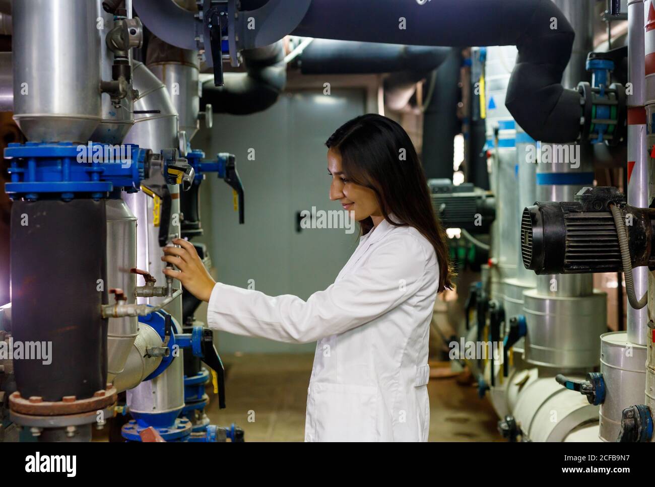 Side view of positive young lady in lab coat checking valves on piping system in industrial facility Stock Photo
