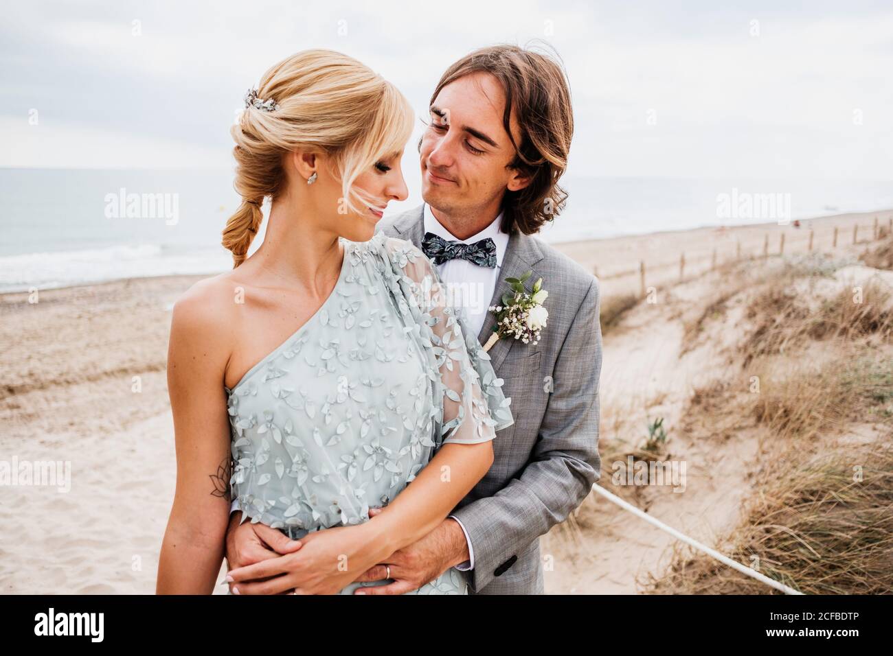 Pleased young groom in wedding suit hugging blonde haired bride in stylish dress behind at empty sandy seashore Stock Photo