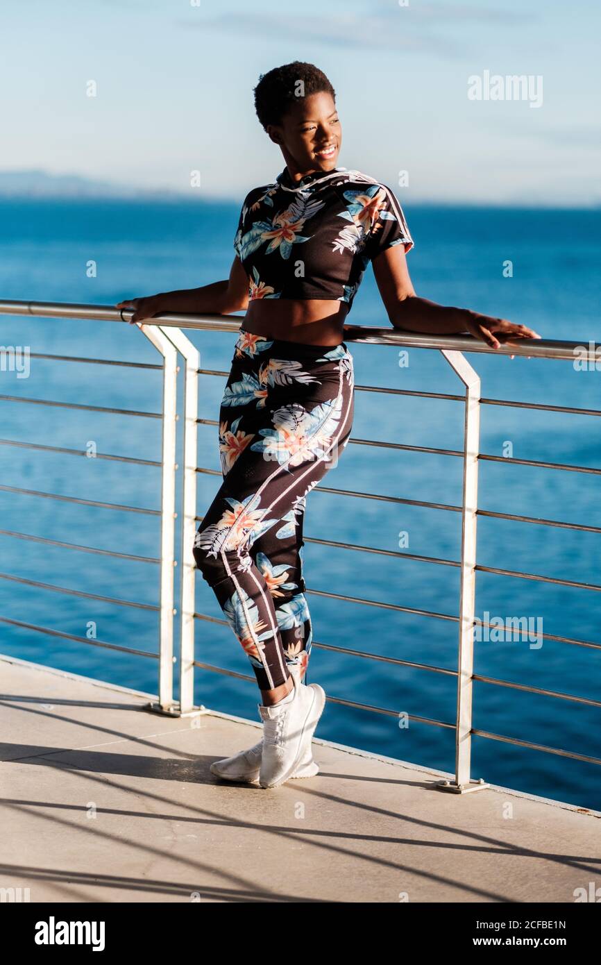 Low angle of charming confident African American lady in flowered sportswear and white sneakers looking away with interest and enjoying life while standing leaning on fence in sunbeams against blue calm sea water Stock Photo