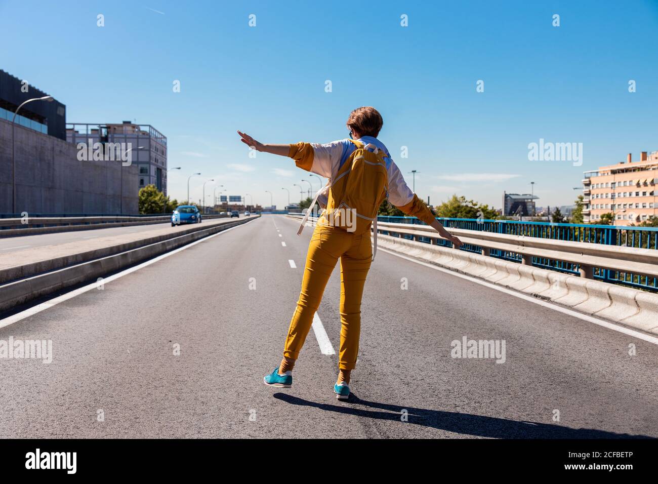 Back view of courageous confident lady in trendy yellow clothes and blue shoes with backpack balancing while walking along marking on city street in bright sunny day Stock Photo