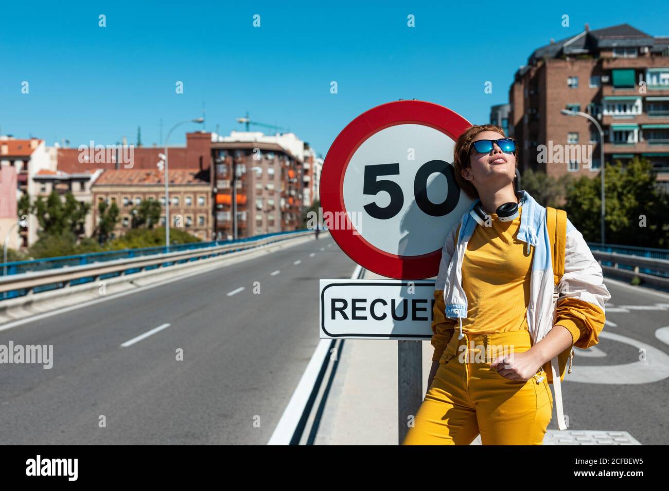 Stylish happy pensive female in sunglasses and yellow clothes with wireless headphones contemplating and smiling while standing beside restrictive traffic sign and looking up against blurred empty road bridge and multistory buildings Stock Photo