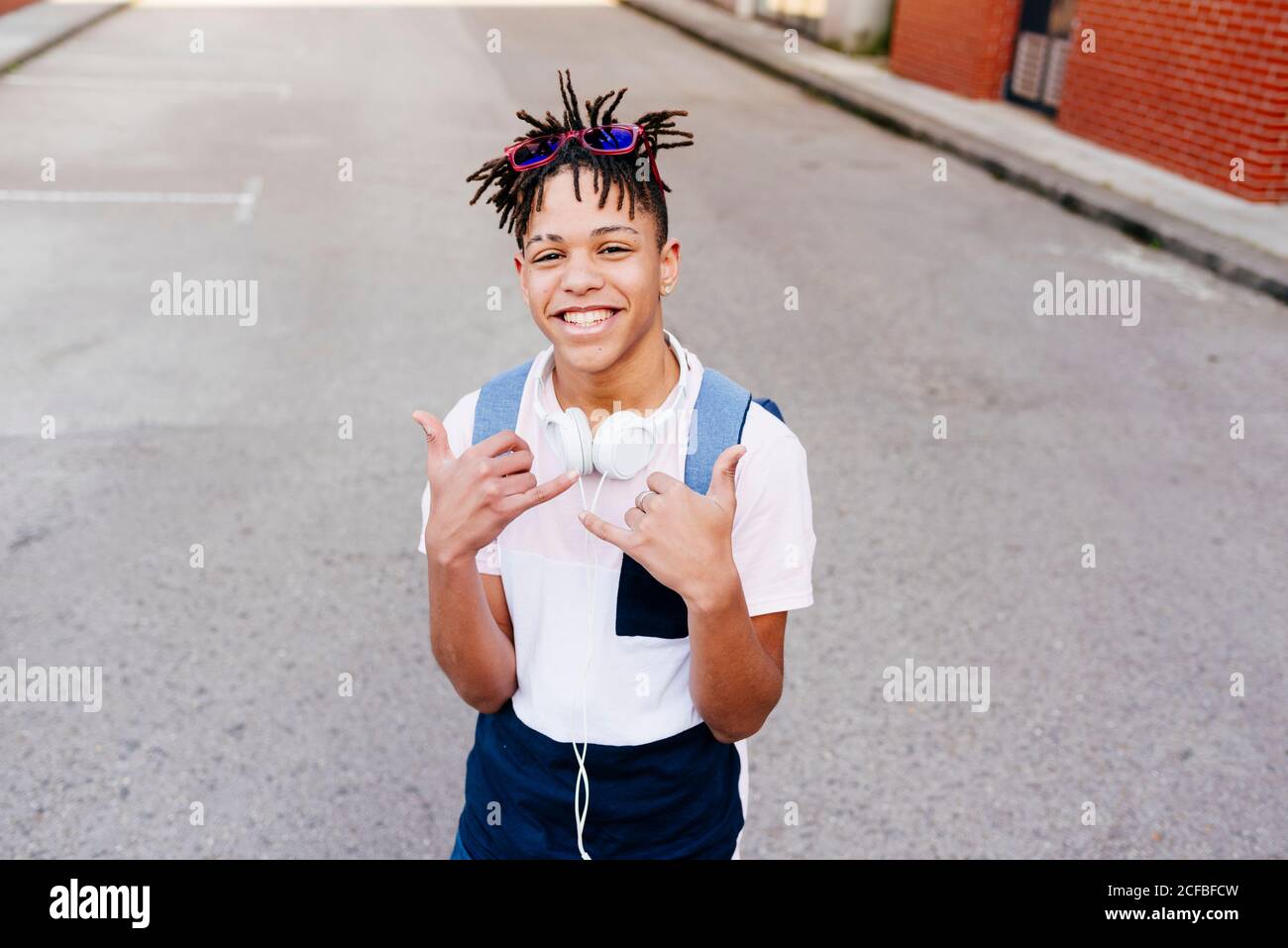 Young joyful African American handsome man in casual apparel with headphones standing on street and showing shaka hand sign Stock Photo