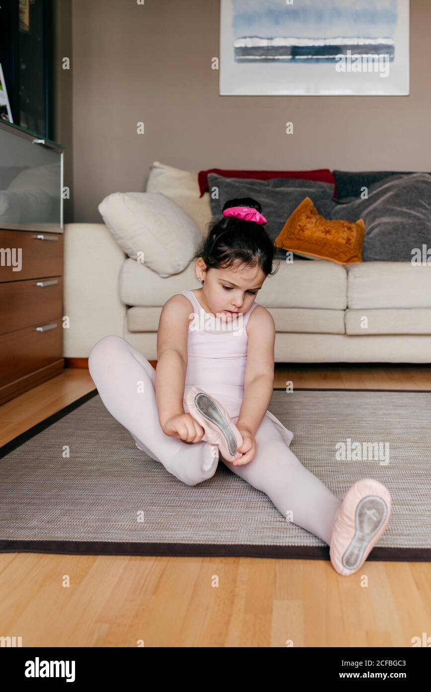 Cute girl in leotard and tights sitting on floor near sofa and putting on  dance shoes before ballet rehearsal at home Stock Photo - Alamy