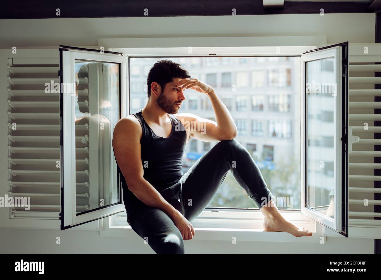 Young successful male in casual singlet sitting on window and looking away Stock Photo