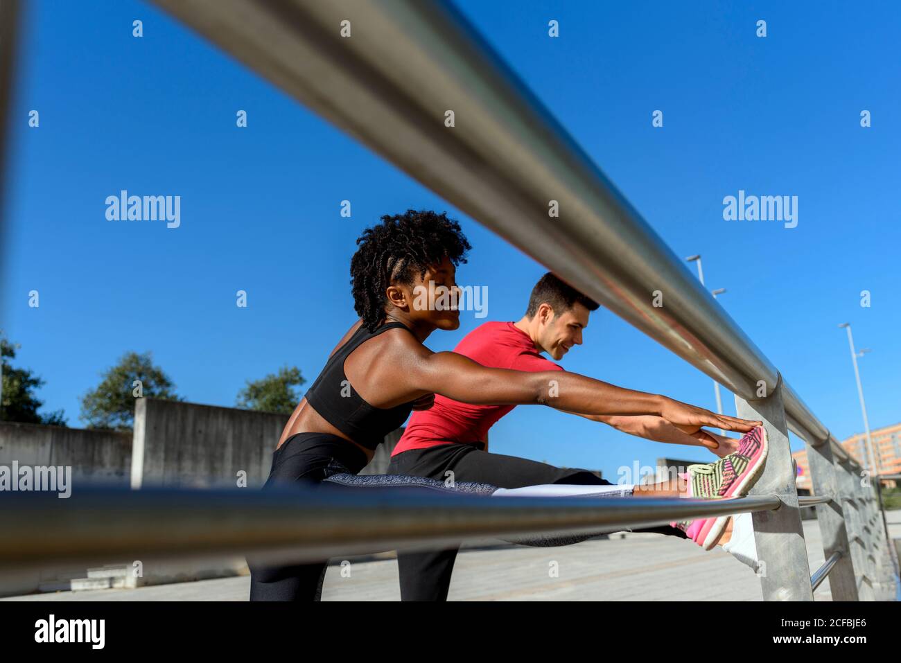 From below side view of cheerful African American fitness woman and smiling sportive man standing next to metal fence and stretching legs while training together on sunny day with blue sky in background Stock Photo