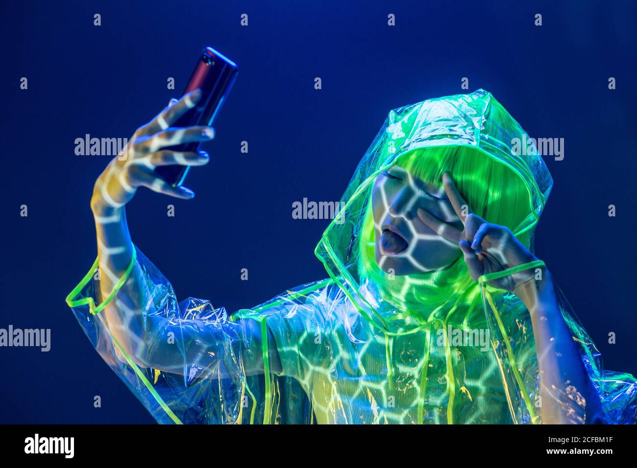 Young Asian woman in futuristic wear and green wig taking selfie on smartphone in fluorescent light Stock Photo