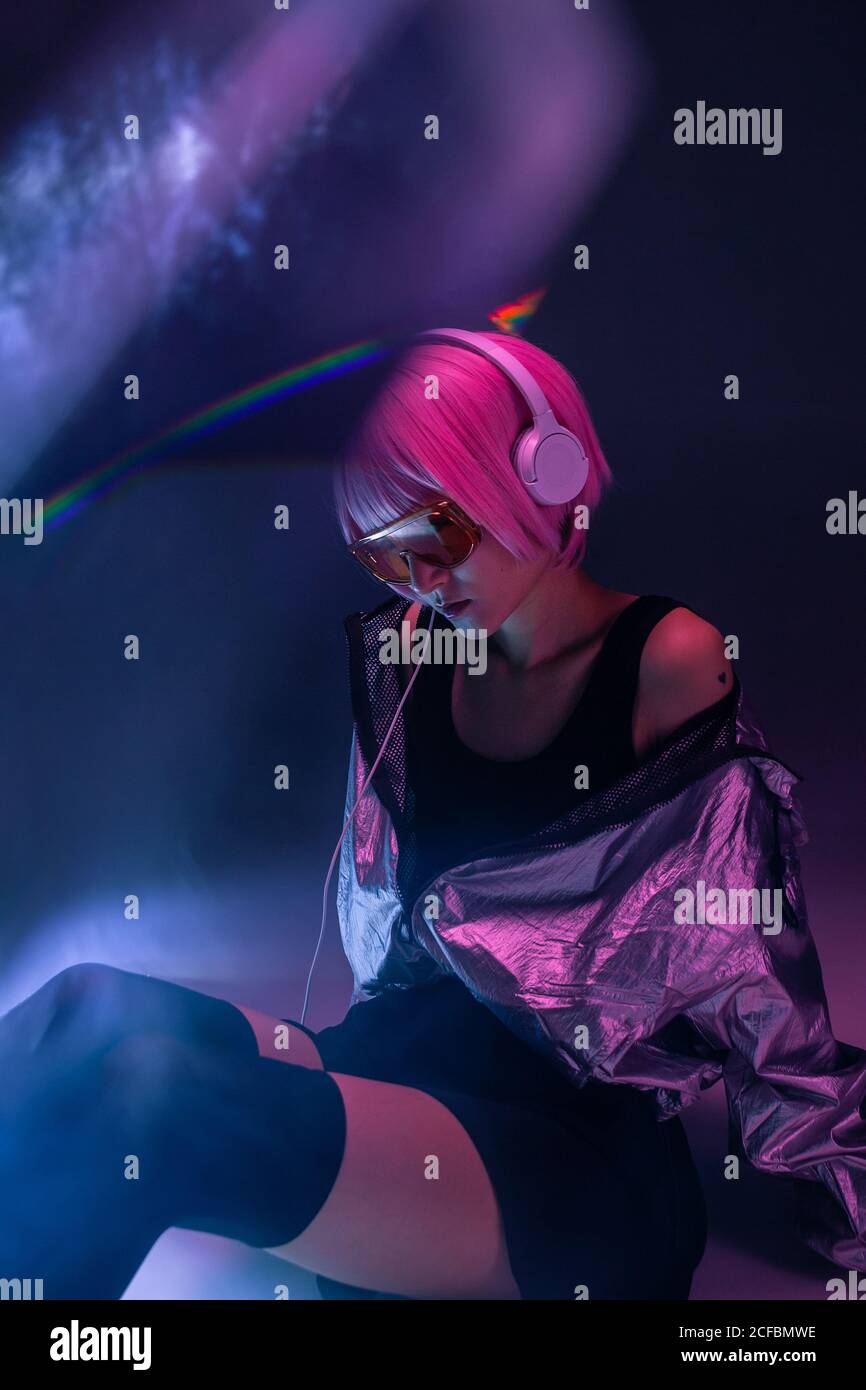 Young stylish Asian Woman wearing in silver jacket with purple hair and headphones sitting on floor while listening to music Stock Photo