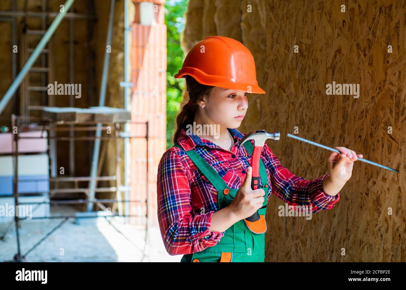 girl hammering a nail. carpentry and woodworking concept. teen child using  hammer tool. building and repair tools. repairman. small girl repairing in  workshop. cheerful kid in helmet hold hammer Stock Photo -