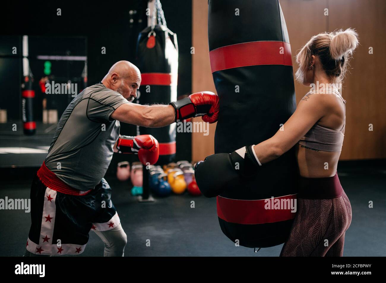 Side view of strong female athlete in boxing gloves holding punching bag  while man practicing punches during workout together in modern boxing gym  Stock Photo - Alamy