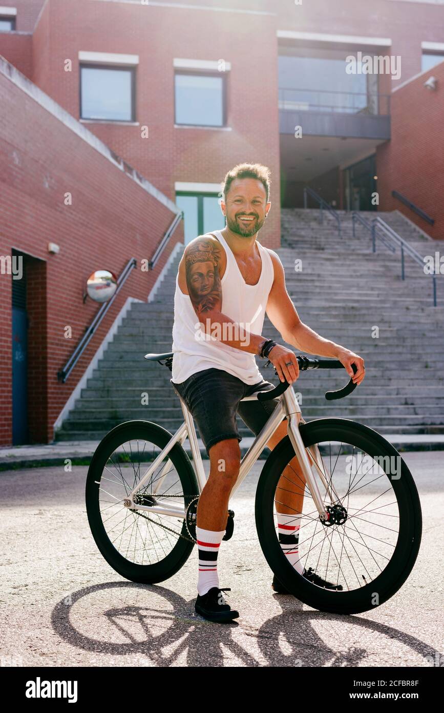 Sportive male wearing white sleeveless shirt and black shorts sitting on bicycle between buildings near stairs on summer sunny day Stock Photo