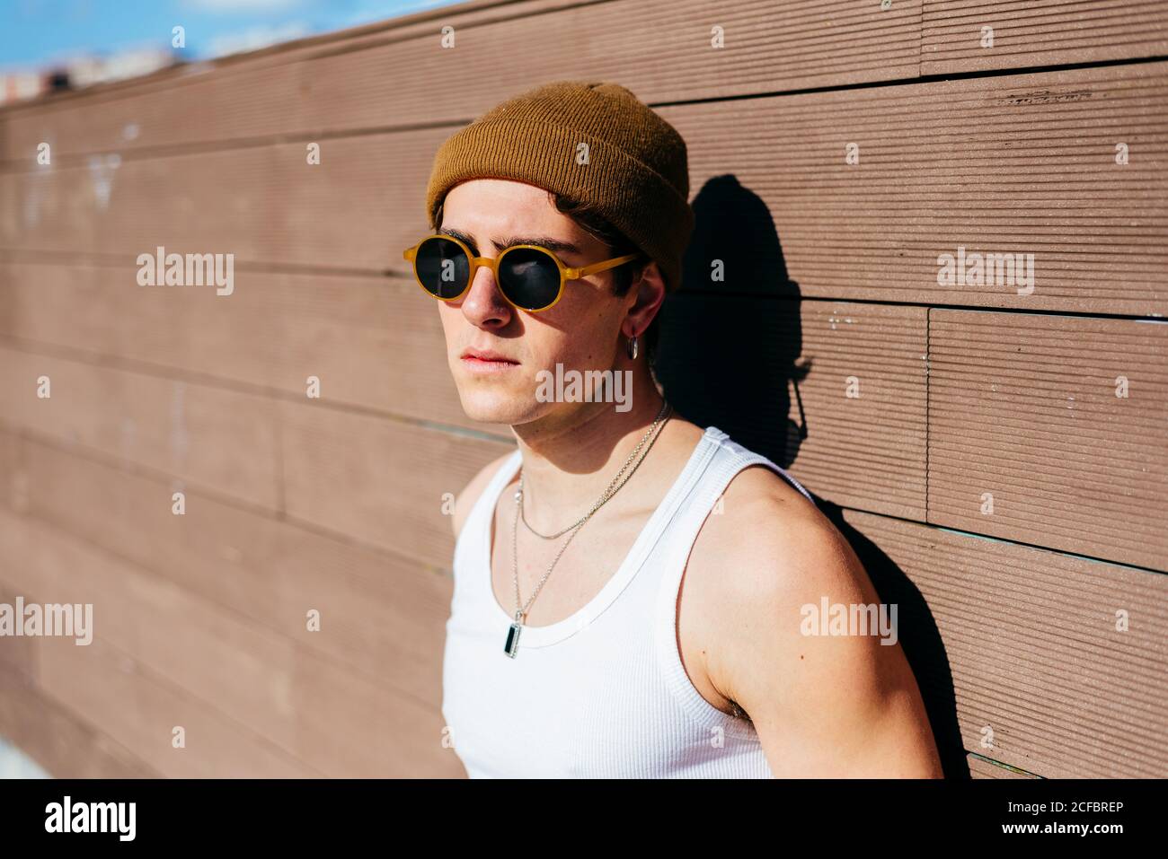 Modern young handsome man in trendy sunglasses and beanie hat and white tank  top standing near brown wall on sunny day on city street Stock Photo - Alamy