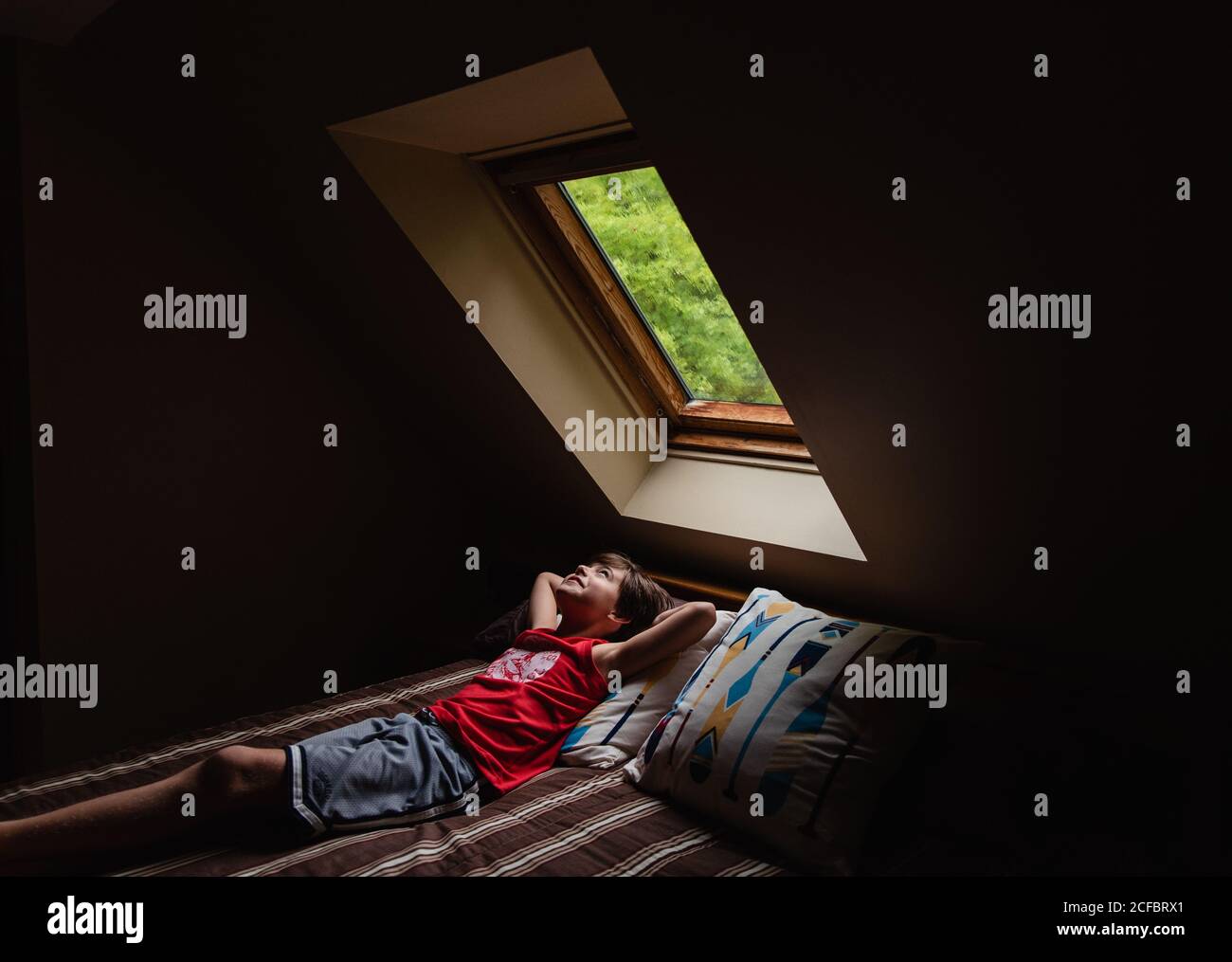 Young boy laying on bed looking up through a sky light in a dark room. Stock Photo