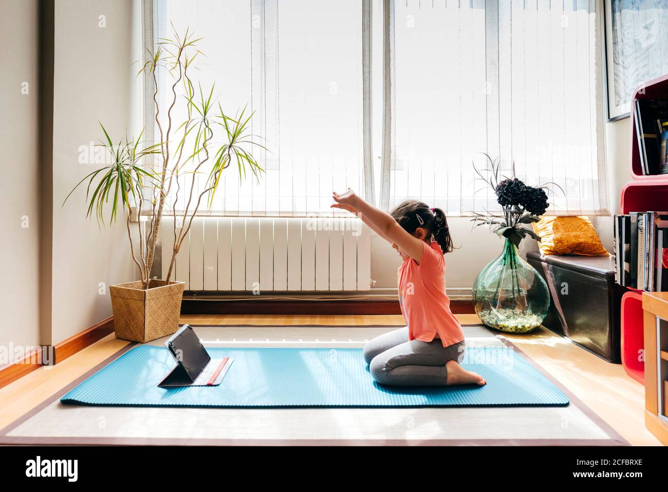 Side view of positive little girl sitting on her knees in yoga pose on mat near window at home while practicing yoga and watching video tutorials on tablet Stock Photo